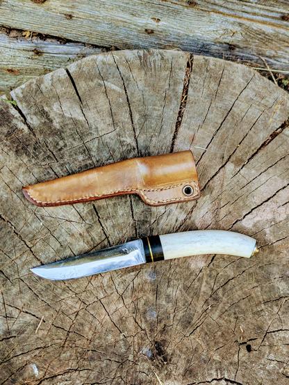 Le couteau et son étui en cuir naturel, posés sur un billot de bois. Vue de dessus 
Le manche est noir et blanc, légèrement incurvé
