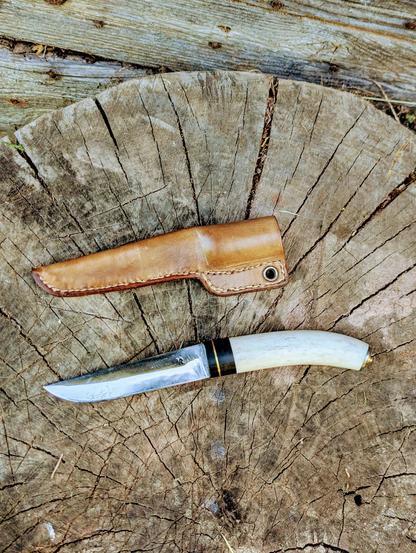 Le couteau et son étui en cuir naturel, posés sur un billot de bois. Vue de dessus 
Le manche est noir et blanc, légèrement incurvé