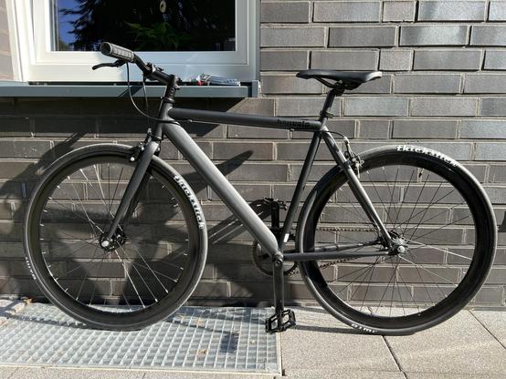 Das Bild zeigt eine moderne, matte Grau-Fixed-Gear-Bicycle, die an einer grauen Backsteinwand gelehnt steht. Die Fahrradrahmen ist schlicht und elegant gestaltet, mit dem Markennamen "bonvelo" deutlich sichtbar. Die Slick-Räder sind schmal. Die Fahrradkette ist sichtbar und verbindet die Pedale mit dem Zahnrad. Der Sattel ist schwarz und passt zur Gesamtkonstruktion. Die Fahrradkette ist sichtbar und verbindet die Pedale mit dem Zahnrad.