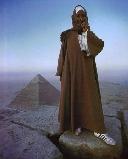 Fotografia de un hombre muy cool con vestimenta típica con tenis addidas fumando un cigarro delante de una piramide en egipto