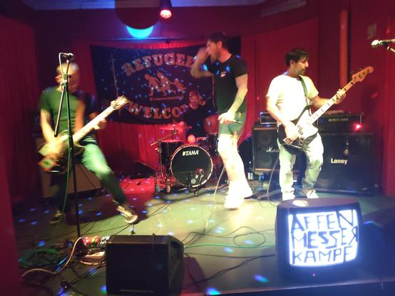 Punk Hardcore Band Affenmesserkampf from Kiel performing live on stage at the Tikozigalpa. There is a huge backdrop on stage that reads "refugees welcome". In front of the stage is an old TV which displays only white noise but has the bands name taped to it.