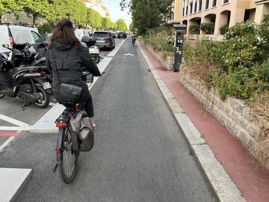 une femme à vélo de dos sur une piste cyclable, des véhicules stationnés à gauche, des immeubles à droite, au fond dans la perspective un autre cycliste