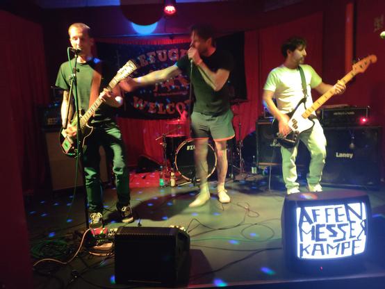 Punk Hardcore Band Affenmesserkampf from Kiel performing live on stage at the Tikozigalpa. There is a huge backdrop on stage that reads "refugees welcome". In front of the stage is an old TV which displays only white noise but has the bands name taped to it.
