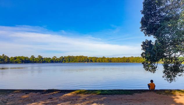 Photo shows a large rectangular reservoir of water mirroring different shades of blue and some green from the surrounding trees. Nearest the photographer there are some red dirt leading up to a stone setting bordering the reservoir. There is a young boy in orange shirt sitting there, looking at the peaceful water. He sits in the shadows of a tree to the right framing the photo. Around the reservoir there is a thick forest with some taller trees. Near the middle of the water there is a small Hindu tower with a spire. The sky is mostly blue, with some lightly clouds to the south/left side.