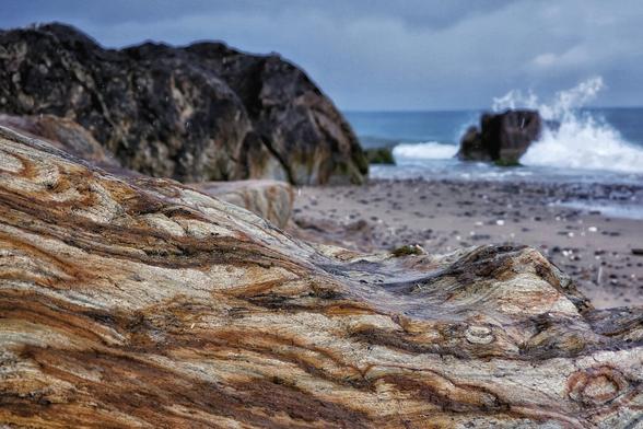 A close-up of a large, textured, multi-coloured rock with swirling patterns, wet from recent rain, dominates the foreground. In the blurred background, a dark, rocky coastline meets a turbulent sea under an overcast sky, with a dramatic wave crashing against a distant rock formation.