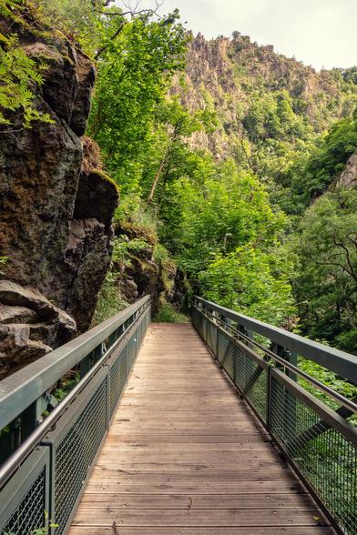 Blick auf eine Holzbrücke. Im Hintergrund das Brücken Ende und dahinter Wald und Berge. Links ragt eine Felswand genau an der Brücke empor.