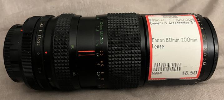 Long black lens with white label dated 8/10/2016 (Canon 80mm-200mm lense, $8.50).

The lens has control rings for zoom, focus, aperture, and rear lens cap release.