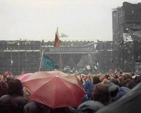 A view over a crowd towards a stage at an outdoor festival. The weather is grey and rainy. Many of the crowd have umbrellas up in a vain attempt to stay dry.