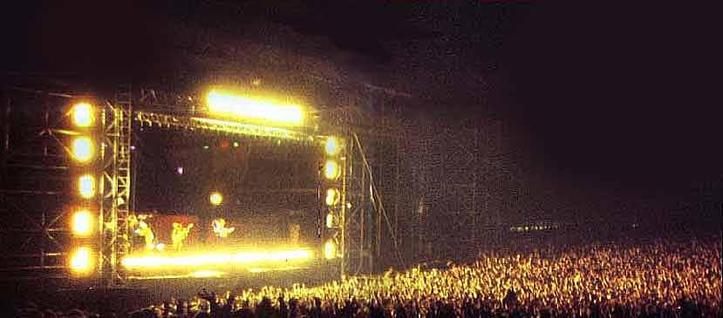 A stage at an outdoor festival after dark. The stage is lit up and the stage lights are spilling out over the front of a large crowd.