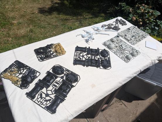 A set of primed sprues laid out on a dust sheet on a garden table: Some fallen statues scatter terrain in black and desert sands, 2 plasma generatorium in black and a nurgle Miasmic Malignifier in rotting flesh. There's also some cranes and machinery in white and steel.