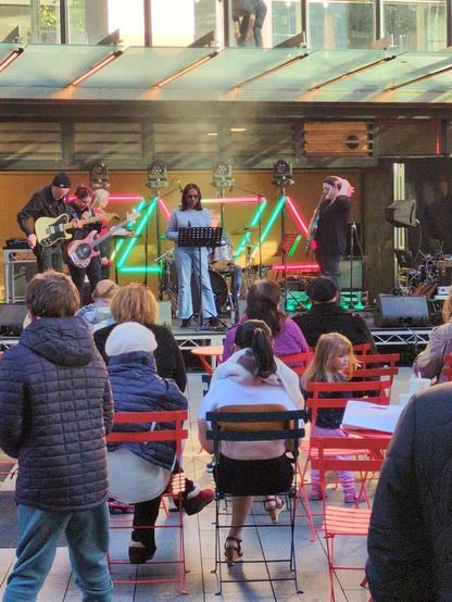 People sitting in garden chairs as local music students perform a rock song in a free public concert. Behind the stage are green and red lights in the shape of triangles.