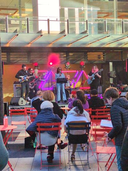 The triangle lights are now purple. A girl in a grey top and sunglasses is singing. Joining her on stage is a lady with pink hair and a blue guitar, a woman dressed in black with shades playing a red bass guitar, a man with a black beanie and a black shirt playing another guitar, and an older gentleman (presumably a music teacher?) on drums.