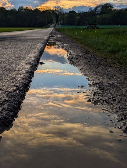 A serene, low-angle shot captures a long puddle along the side of a country road at dusk. The sky, painted with soft orange and grey clouds, is perfectly reflected in the still water of the puddle, creating a mirror image. The tarmac road stretches into the distance on the left, while a grassy verge and a field, with hints of agricultural machinery, line the right. A dark band of trees forms the horizon, silhouetted against the colourful twilight sky.