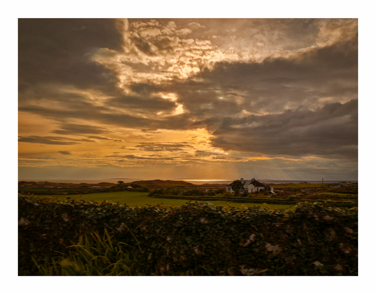 The photo is of a picturesque landscape at sunset.  There is a dramatic sky filled with golden and dark gray clouds which takes up the upper two-thirds of the frame.  There is a single, white house nestled in a green field in the mid ground, near the horizon.  In front of the house and field, a dense hedge stretches across the photograph, partially obscuring the lower portion.