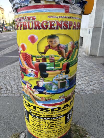 Buntes Werbeplakat an einem Laternenpfahl für ein Hüpfburgenfest in Potsdam mit fröhlichen Kinderbildern, Infos zu Öffnungszeiten und dem Standort. Einzelne Kleinkinder sind auf dem Plakat zu sehen, jedes in einer Art Blase sitzend und liegend illustriert.