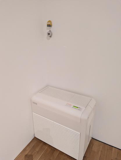 Corner of a room, white walls wood parquet flooring. In the corner are two dataloggers (small plastic boxes) one above the other, fixed to the wall. Directly below them is a running portable air conditioning unit