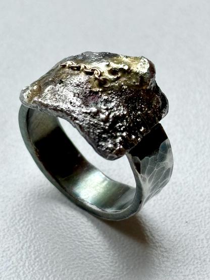 A unique ring featuring a textured, metallic top with a rough finish and a small gold chain detail. The band is hammered and polished, contrasting with the rugged top. The background is a simple, light color.