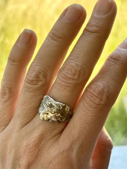 A close-up of a hand wearing a textured silver ring. The background features soft green and yellow tones, suggesting a natural outdoor setting.