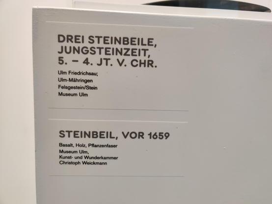 The text label from the exhibit. It reads, in German, "three axe heads, early stone age, fourth to fifth millennium before Christ" and "Axe head, before 1659".
