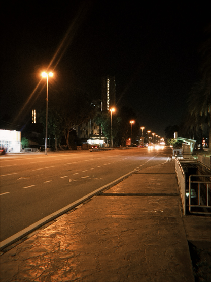 The image depicts a nighttime urban street scene. The street is illuminated by streetlights, casting a warm glow on the asphalt. The road is marked with white dashed lines and arrows, indicating lanes and directions. On the left side, there is a sidewalk with a textured surface, and on the right, a metal railing is visible, possibly indicating a pedestrian crossing or a barrier. In the background, a tall building with illuminated windows stands out against the dark sky, suggesting a city environment. The street is relatively empty, with a few vehicles visible in the distance, their headlights and taillights creating streaks of light. Trees line the street, adding greenery to the urban setting. The overall atmosphere is calm and quiet, typical of a city at night.