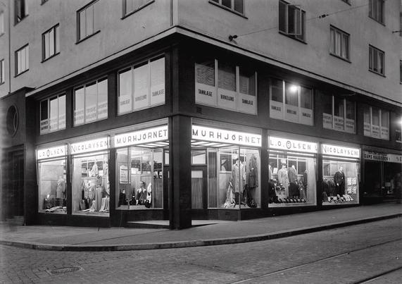 A black and white photo of a corner store in a 30s brick building. Dark walls lit by fashionable bright lights. Store front reading Murhjørnet and L.O. Olsen & Nicolaysen A/S, and signage for a dentist in the second floor.