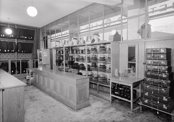 A black and white photo of a menswear shop, showing hats, suits, gloves, and briefcases on display.