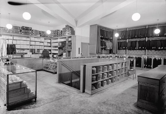 A black and white photo of a menswear shop, showing suits, cufflinks, flatcap hats, socks and other items of clothing. A stair leading down to the floor beneath.