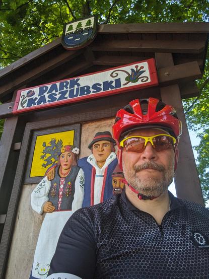 Selfie w czarnej koszulce w białe sześciokąty, czerwonym kasku i żółtych okularach rowerowych. Za mną napis "Park kaszubski" i drewniana płaskorzeźba pary w ludowych strojach.