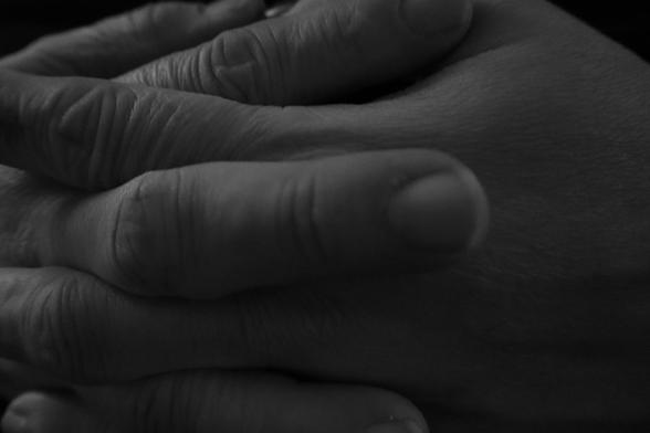 This photograph shows a close-up of two hands gently clasped together, with fingers resting softly across the back of the other hand. The lighting is low, creating deep shadows and emphasizing the texture of the skin and the lines on the fingers. The image is in black and white, enhancing the contrast and intimacy of the composition.