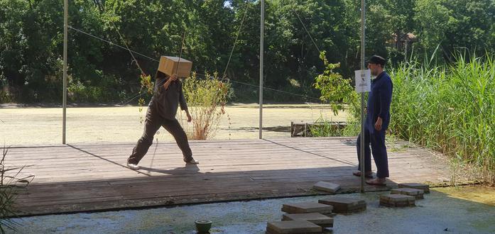 Two people on a wooden platform with trees and scummy green water behind. One is playing nylon strings that attach to a suspended cardboard box with a violin bow