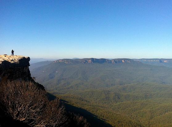 A lookout over a sunny canyon. The lookout is the edge of an rocky cliff that drops steeply into the canyon. Two small figures stand close to each other looking out at the view, into the distance. There is no safety fence. In the distance are more straight sided rock cliffs overlooking the canyon. The sky is clear above.