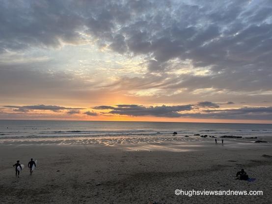 A photo of the sunset on Fistral Beach in Newquay, England. On the beach, some surfers are walking towards the sea.