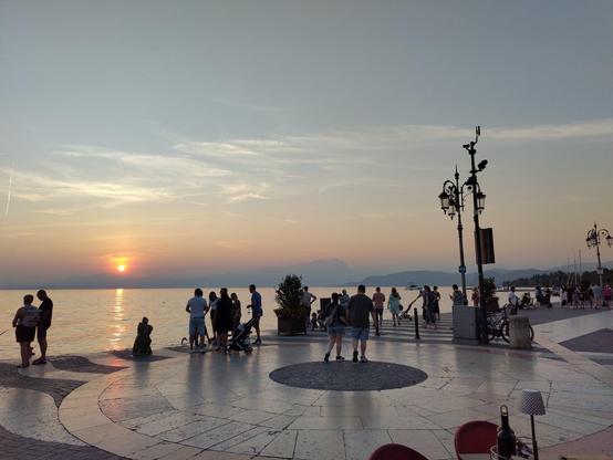 Blick über gepflasterten Platz mit einigen Leuten.
Im Hintergrund der See und die Sonne.
Die Sonne ist bereits so tief, dass sie gleich hinter'm Horizont verschwindet und nur noch ein nicht blendender orangener Ball ist