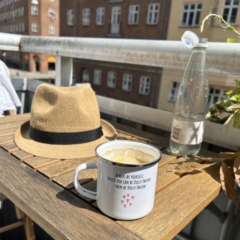 Hat, water bottle and coffee cup reading "Always be yourself unless you can be Dolly Parton - then be Dolly Parton" on a small balcony table bathed in sun, some buildings in the background