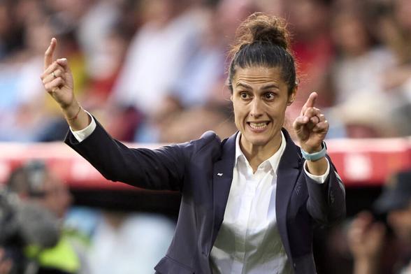 Montse Tomé in Hemd und Jackett von der Hüfte aufwärts am Spielfeldrand, sie hat beide Arme ungefähr auf Kopfhöhe und beide Zeigefinger ausgestreckt, gibt ihren Spielerinnen so Anweisungen. Ihre Stirn ist beim Sprechen gerunzelt, ihr dunkles Haar trägt sie in einem Dutt.