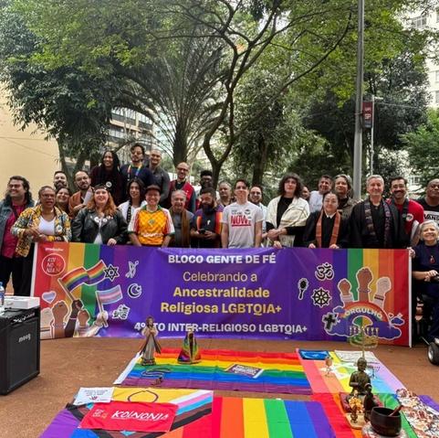 People of all religions, races, ages, and genders stand proudly behind a Portuguese-language banner celebrating LGBTQIA+ religious ancestors. On the right side are two Jodo Shinshu clergy, in black robes. Before the banner is a tableau created from various rainbow flags with some religious items, most prominently two buddhas statues with a Buddhist bell. Photo is from Rev. Jean Tetsuji.