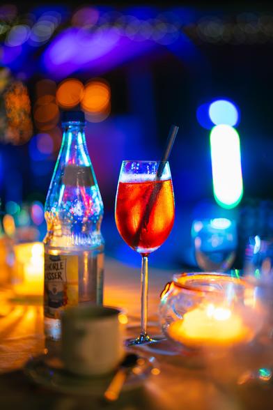 A vibrant cocktail sits elegantly in a tall glass with a black straw, glowing orange under ambient lights. Beside it, a sparkling water bottle reflects multicoloured lights, adding shimmer. The background features blurred blue and orange bokeh, creating a lively, festive atmosphere. Nearby, a lit candle in a glass holder casts a warm, inviting glow on the table.