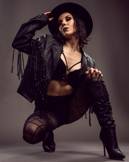Person wearing a black leather jacket with fringe, a black bra, and high-waisted shorts. Holds a wide-brimmed black hat while kneeling, with crossed black tights and knee-high heeled boots. Smoky eye makeup and dark lipstick accentuate the outfit. Neutral gray background.