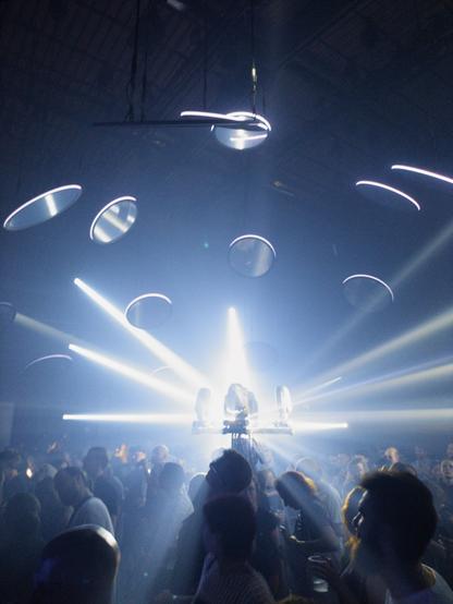 Multiple beam emitters sit in the center of a crowd of people, shooting bright blue light into round mirrors hovering above the crowd.