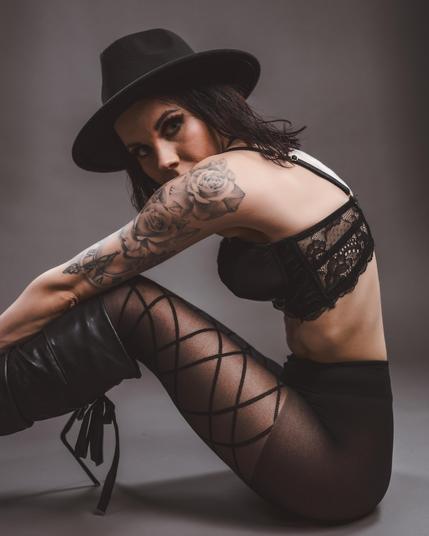 Woman sitting on the floor wearing a black hat, black lace bra, and black boots with laces. Her arms and legs are crossed, with a tattoo of roses on her left arm. She wears sheer stockings with a crisscross pattern. The background is a neutral gray.