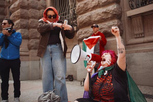 We can see one protester on ground sitting and doing slogans on megaphone, one photographer at the edge of the left side of the photo and someone in between with Palestine cola on hand. At the background we can see one more person holding Palestine flag.