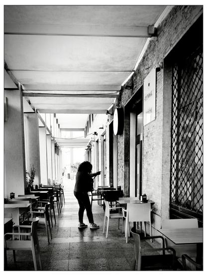 Immagine in bianco e nero di un porticato, una donna dai capelli ricci cammina tra i tavolini vuoti di un bar all’aperto. Ha una mano infilata in una borsa, l’altra sembra in movimento, come se stesse sistemando qualcosa o cercando un oggetto. L’ambiente è urbano ma silenzioso, i tavoli e le sedie sono ordinati, e la luce naturale filtra delicatamente dal fondo del porticato.