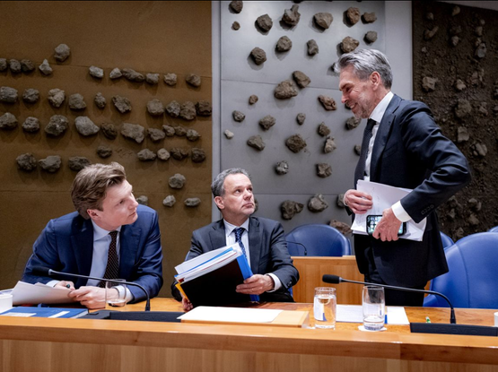 Foto van de tweede kamer met drie heren in blauw pak achter een tafel, met op de achtergrond een muur in drie vlakken met op ieder vlak een aantal grote stenen bevestigd in een willekeurig patroon.