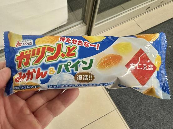ガツンとみかん&パイン杏仁豆腐アイスバーのパッケージを手に持っている。パッケージは青地に白い杏仁豆腐アイスのイラスト。みかんやパインも描かれている。