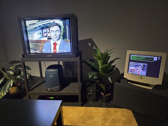 Eine Art Wohnzimmeraufstellung mit Couchtisch und Pflanzen mit einem Röhren-TV, auf dem eine interaktive Installation läuft. Auf einem CRT daneben sieht man den Irix-Desktop. Unter dem TV steht eine Irix O2, ein schwarz-blauer Tower mit geschwungenem Gehäuse. Die Workstation ist mit Strom und den Monitoren verbunden.