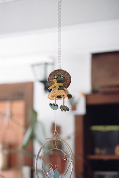 Marionnette d'une grenouille avec un chapeau de paille, semblant porter le poids de son destin par le pied. Elle semble tout à fait heureuse malgré tout.