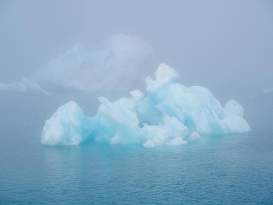 A piece of blueish iceberg that's shaped almost like a cloud, floating on a glacier lagoon in the mist. There are a couple other floating pieces of icebergs in the background, obscured by the mist.