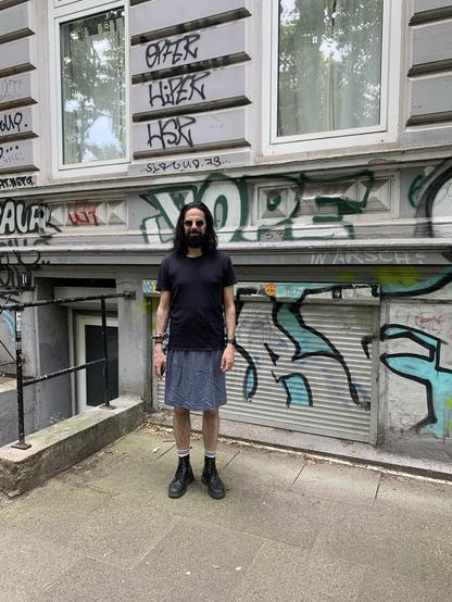 A picture of me, a man in his late thirties with shoulder-long black, lightly curled hair. Also, I have a considerable black beard. I am wearing sunglasses. And, which is the whole point of this picture, I am wearing a knee-length blue skirt. Also, I have black Dr Martens boots on. I am standing in front of an apartment building, there is graffiti on the walls.