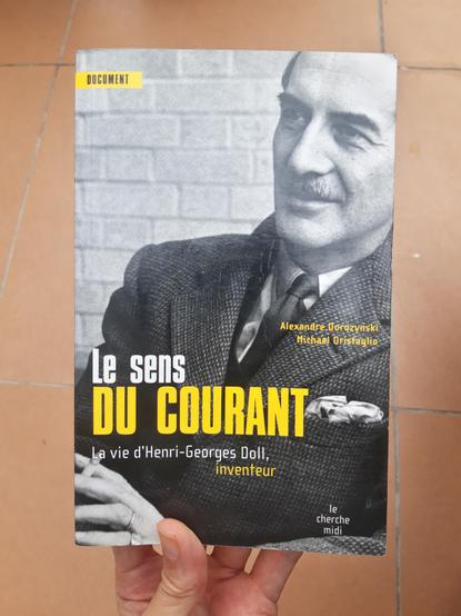 A hand holds up a copy of a book entitled "Le sens du courant, La vie d'Henri-Georges Doll, Inventeur" by Dorozynski and Oristaglio.