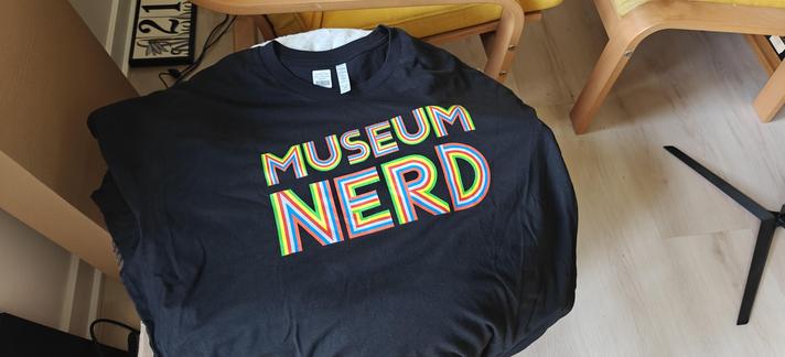 A black T-shirt with the words "Museum Nerd" in uppercase rainbow colored lettering.